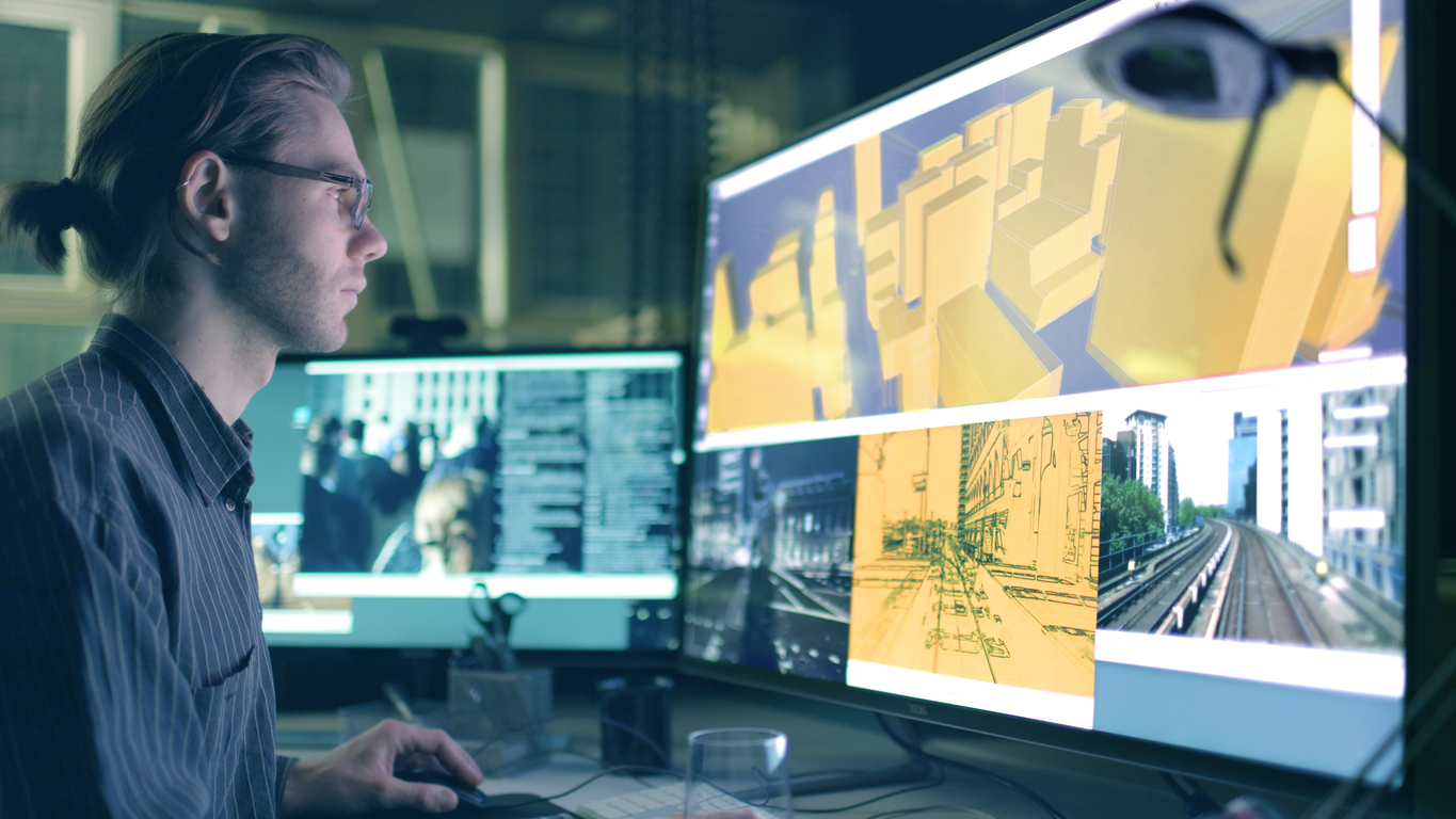 A young man working in a dark office environment using AI-Powered Imagery Intelligence for planning designs on two computer screens.