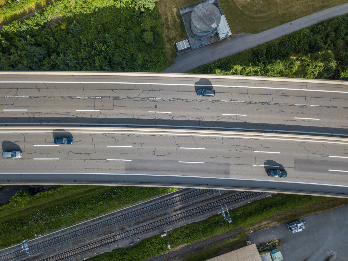 Pothole Lifecycle Management: Aerial view showing a highway with visible cracking on the pavement, passing above some railroad tracks and green trees.
