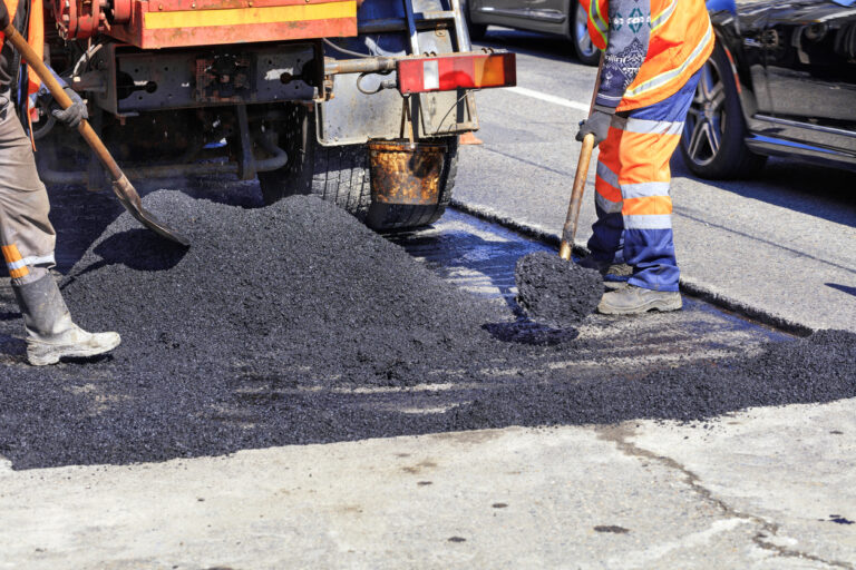 Pothole Lifecycle Management: Two road workers in bright orange safety vests are shoveling a pile of fresh black asphalt onto a road surface next to a truck performing pavement repairs.