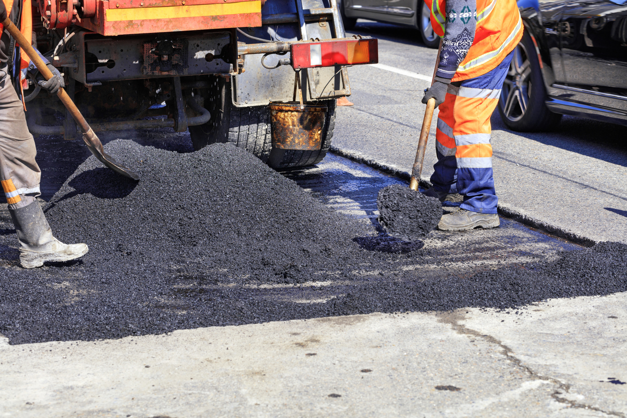 Pothole Lifecycle Management: Two road workers in bright orange safety vests are shoveling a pile of fresh black asphalt onto a road surface next to a truck performing pavement repairs.