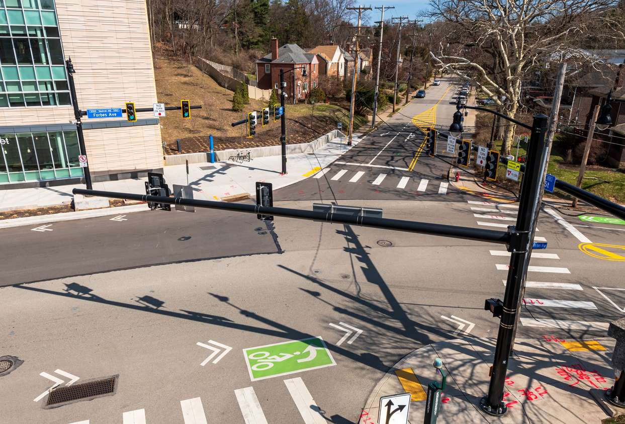 Connected and Autonomous Vehicle Infrastructure: An overhead view of a modern city intersection featuring high-visibility crosswalks, dedicated green bike lanes, and multiple traffic signal poles.