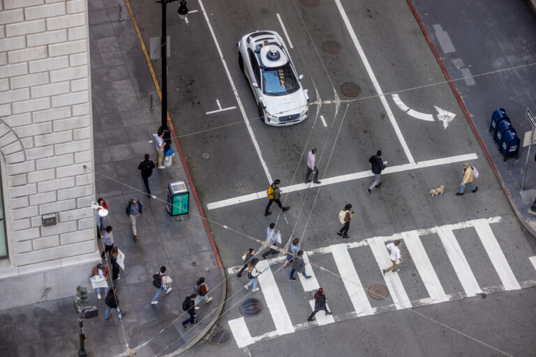 Connected and Autonomous Vehicle Infrastructure: An overhead view of pedestrians crossing a city street as an autonomous vehicle waits at the intersection.