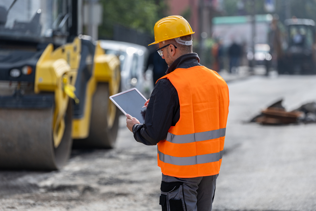 Predictive Pavement Maintenance: A construction worker in a yellow hard hat and orange safety vest stands on a road, using a digital tablet next to a large yellow steamroller.