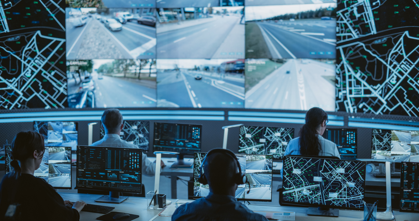Connected and Autonomous Vehicle Infrastructure: Four specialists sit in a high-tech control room, monitoring multiple screens that display live traffic camera feeds and digital city maps.